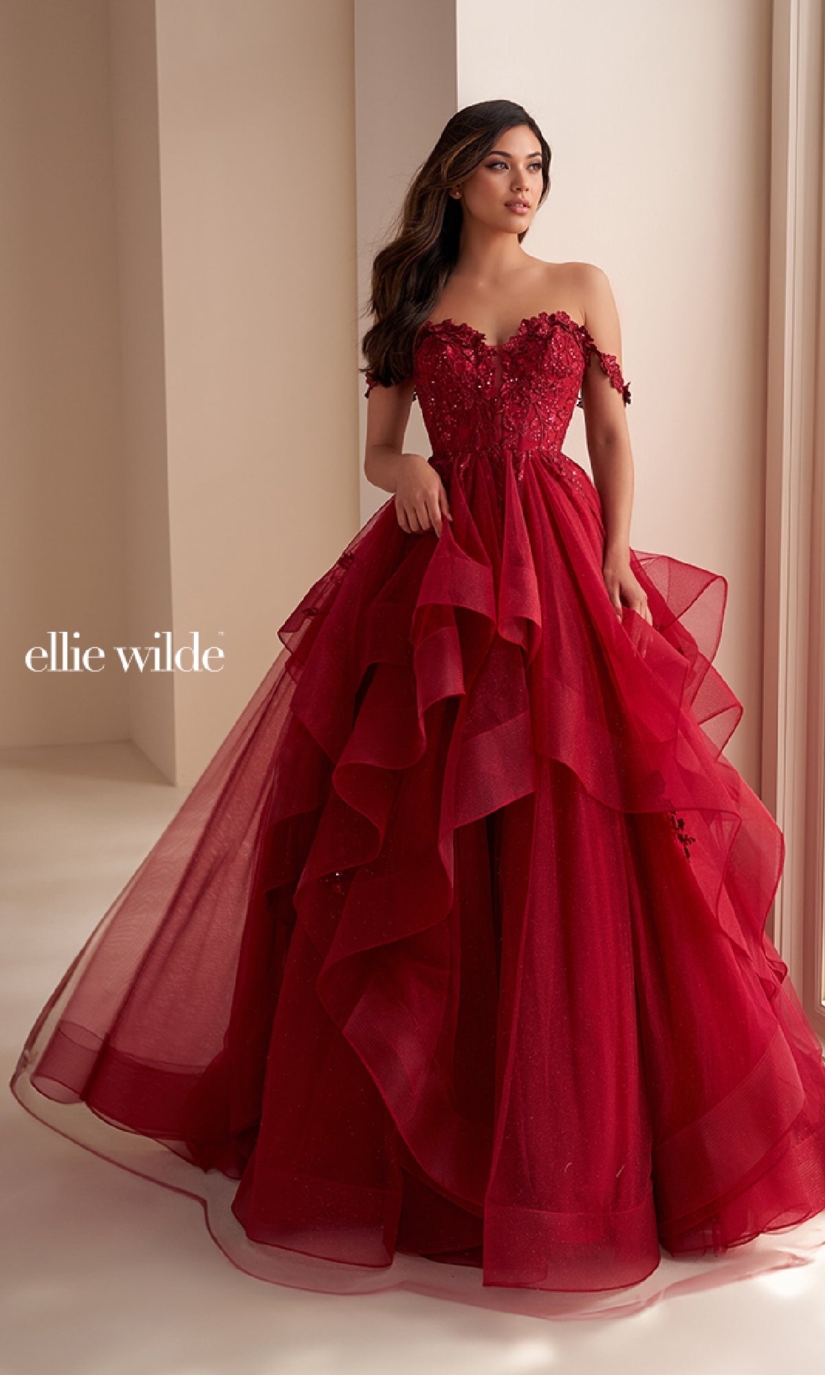 A woman wears the Ellie Wilde Long Designer Prom Ball Gown EW35084— a strapless, layered deep red glitter tulle dress with floral accents and off-the-shoulder sleeves. She stands by an indoor window in this stunning designer gown.